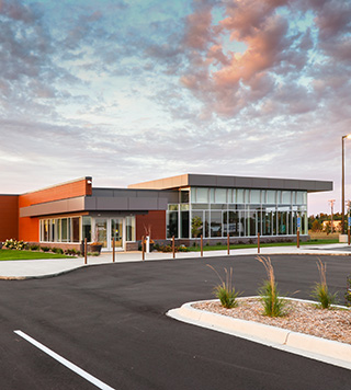 Image of a small clinic with lots of windows. The clinic has a large parking lot with landscape. The sun is setting on a partly cloudy sky.