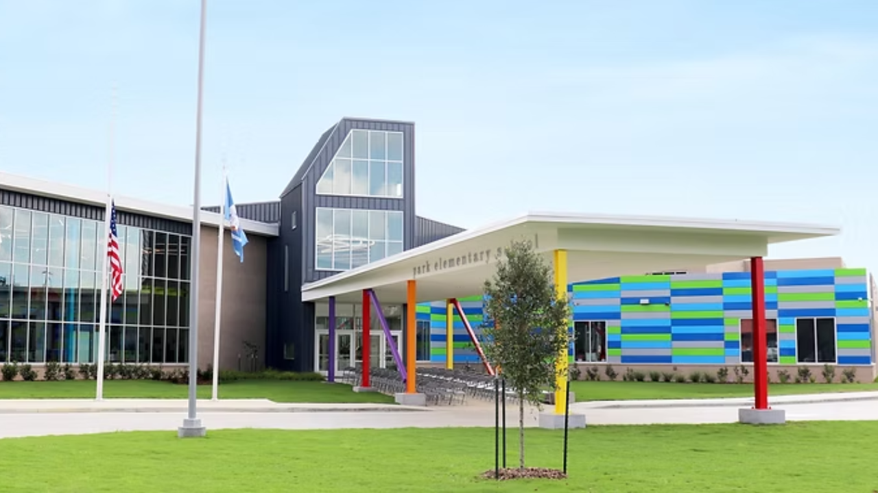 Exterior of a modern elementary school with expansive glass walls, colorful striped facade panels, covered entryway, and landscaped grounds under a clear sky.