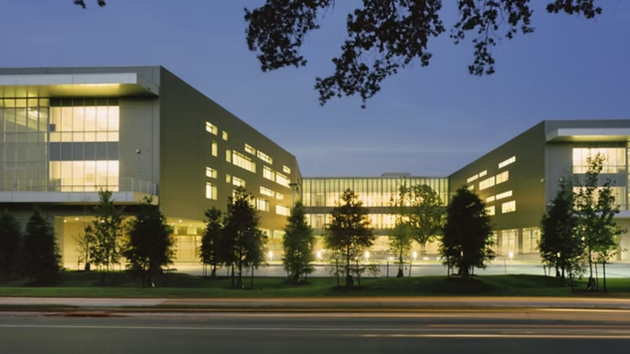 Modern academic or institutional building complex illuminated at dusk with two symmetrical wings, a glass central connector, and trees lining the landscaped courtyard along a roadway.