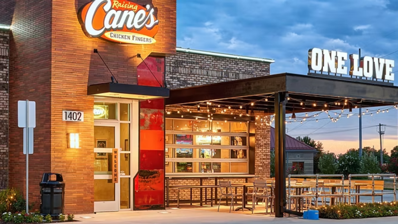 Exterior of a Raising Cane’s Chicken Fingers restaurant at dusk featuring brick architecture, illuminated signage, a covered patio with string lights, and outdoor seating under a One Love canopy.