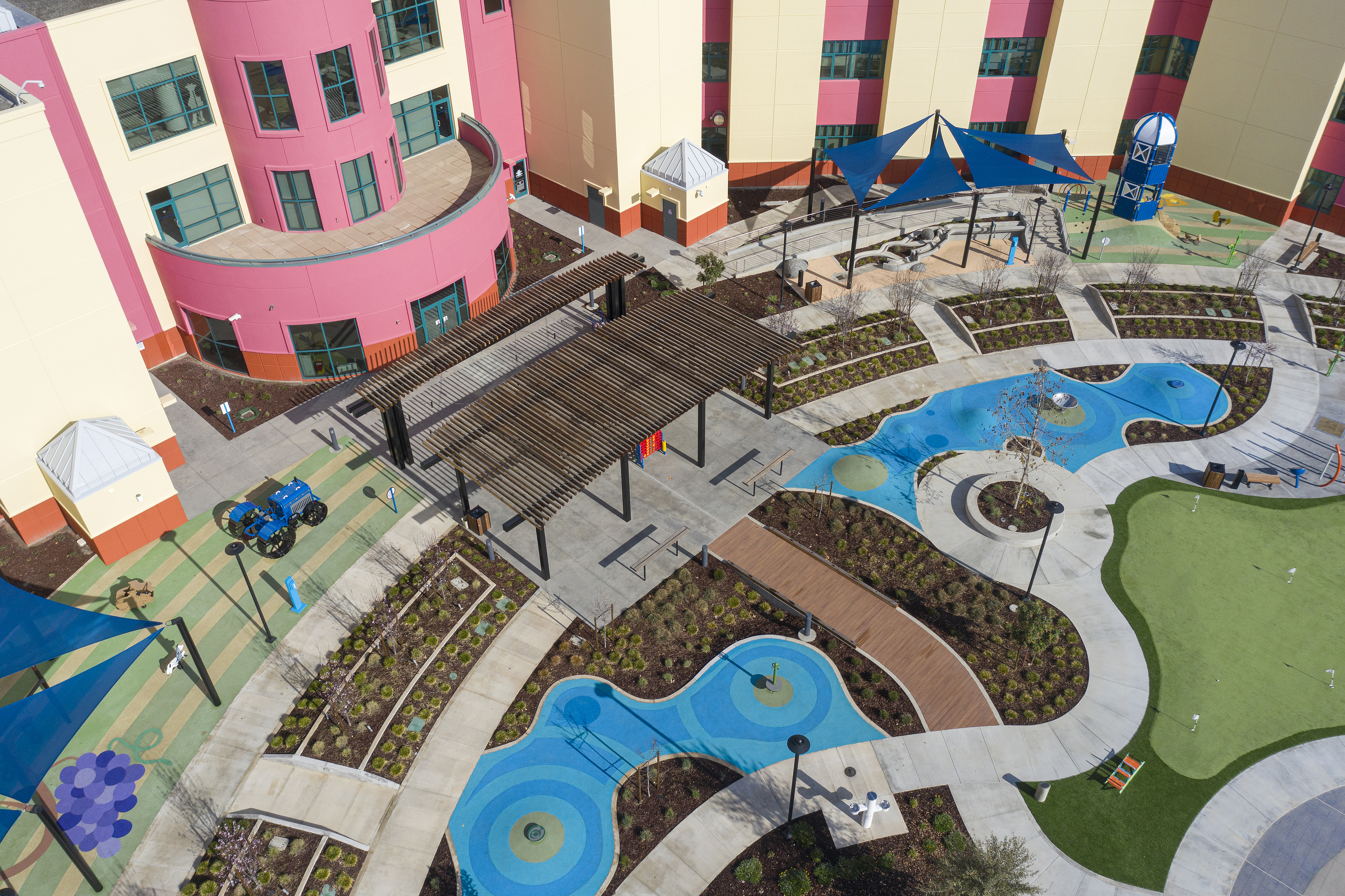Aerial view of a colorful outdoor courtyard with playground equipment, blue shade canopies, landscaped garden beds, walking paths, seating areas, and bright pink and cream buildings.